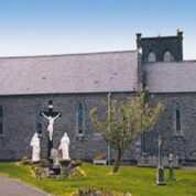 Stone church with arched windows, outdoor crucifix, statues, small garden, and gravestones under a clear blue sky.