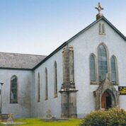 A stone church with arched windows, a cross on the roof, and a grassy area in front under a clear sky.