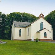 Small cream-coloured church with arched windows, a bell tower, and a statue in front, surrounded by green grass and trees.