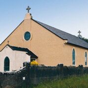 A beige church with arched windows and crosses on the roof, surrounded by a stone wall and grassy area, with a mobile phone mast in the background.