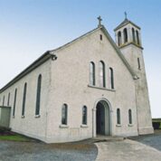 A white church with arched windows and a bell tower stands beside a path, surrounded by grass under a clear sky.