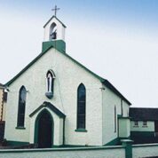 Small white church with green trim, a bell tower, arched windows, and a statue above the entrance, next to neighbouring houses.