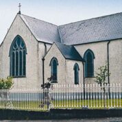 A stone church with arched windows, a slate roof, and a white fence surrounding a green lawn.