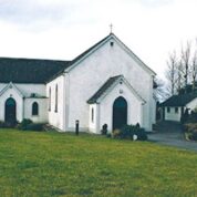 A white church with a dark roof stands on a grassy lawn, with trees and a small building in the background.