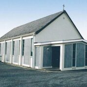 A small, white church with a dark roof and a cross on top, set on a paved car park with a statue visible to the side.