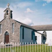 A stone church with arched windows and a small bell tower, surrounded by a white fence under a partly cloudy sky.