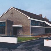 Modern brick church building with large stained glass windows and a sloped roof, surrounded by a low white wall.