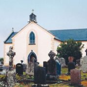 A small church with a blue roof stands behind old gravestones in a rural cemetery, surrounded by trees and greenery.
