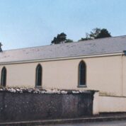 A small cream-coloured church with arched windows, a slate roof, and blue decorative trim, behind a stone wall.