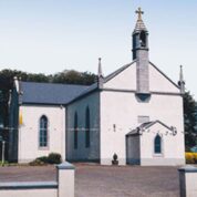 Small white church with a bell tower, cross on top, arched windows, and flags, set against trees and a clear sky.