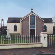 A small church with a cross on top, arched windows, a bell tower, and a fenced yard with a grave and a cross headstone.
