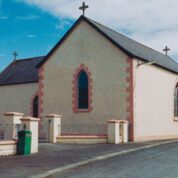 Small beige church with arched windows, stone trim, and crosses on the roof, surrounded by a low fence and a paved road.