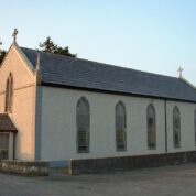 A small, light-coloured church with arched windows, a slate roof, and crosses on the roof peaks, surrounded by a low wall.