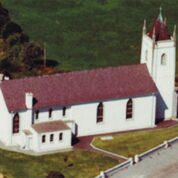 Aerial view of a white church with a red roof and tower, surrounded by green grass and a paved area.