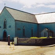 A small, light blue church with arched windows and a slate roof, surrounded by a low wall under a partly cloudy sky.