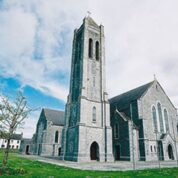 A large stone church with a tall bell tower stands on a green lawn under a partly cloudy sky.