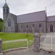 A stone church with a tall bell tower, arched windows, and a gated entrance surrounded by green grass.