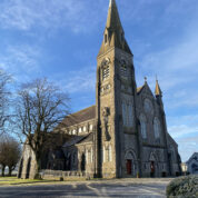 A large stone church with a tall spire, arched windows, and pointed turrets under a clear blue sky.