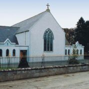 A white church with a large arched window and cross on the roof, surrounded by trees and a stone wall.