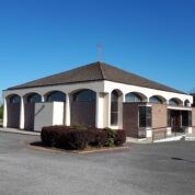 A modern church with a brown tiled roof, arched windows, and a cross on top, set against a clear blue sky.