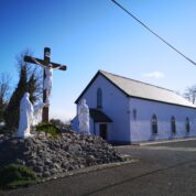 A small white church with a crucifixion scene and statues in front, set under a clear blue sky.