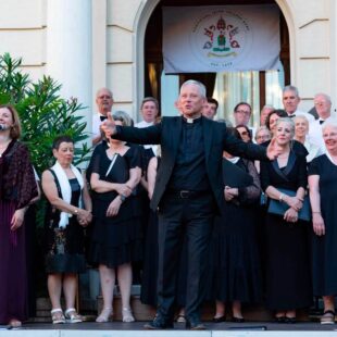 A priest gestures whilst standing with a choir of men and women singing outside a building with pillars.