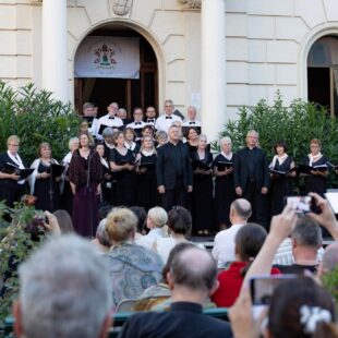 A choir performs outdoors in front of a building whilst an audience watches and someone conducts in the foreground.