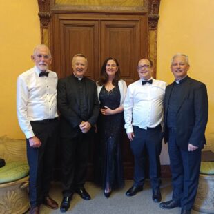 Five people in formal attire stand smiling in front of a large wooden door with ornate trim.