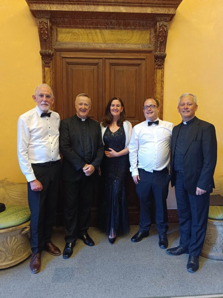 Five people in formal attire stand smiling in front of a large wooden door with ornate trim.