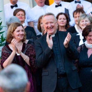 A smiling priest and two women applaud among a large, joyful group dressed in formal black and white attire.
