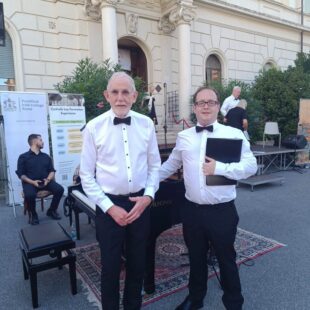 Two men in white shirts and black trousers stand in front of a piano outdoors, with a building and banners behind them.