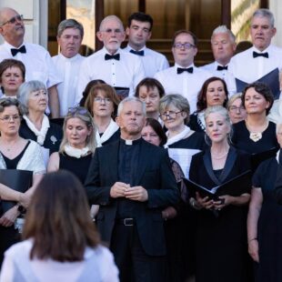A choir of men and women, some in clerical attire, stand on steps outdoors, facing a conductor.