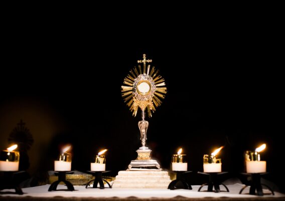 A gold monstrance with the Eucharist, surrounded by six lit candles, stands on an altar against a dark background.