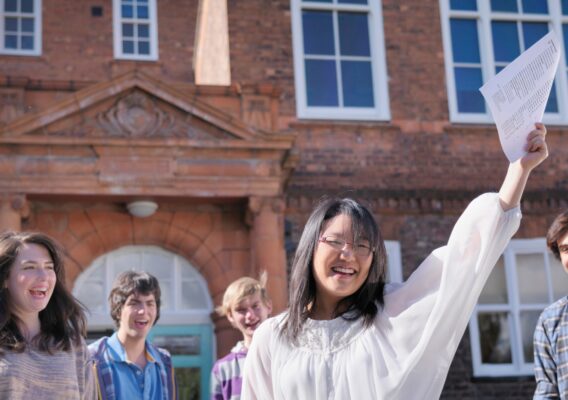 A smiling student holds up a sheet of paper outside a brick building, surrounded by happy friends.