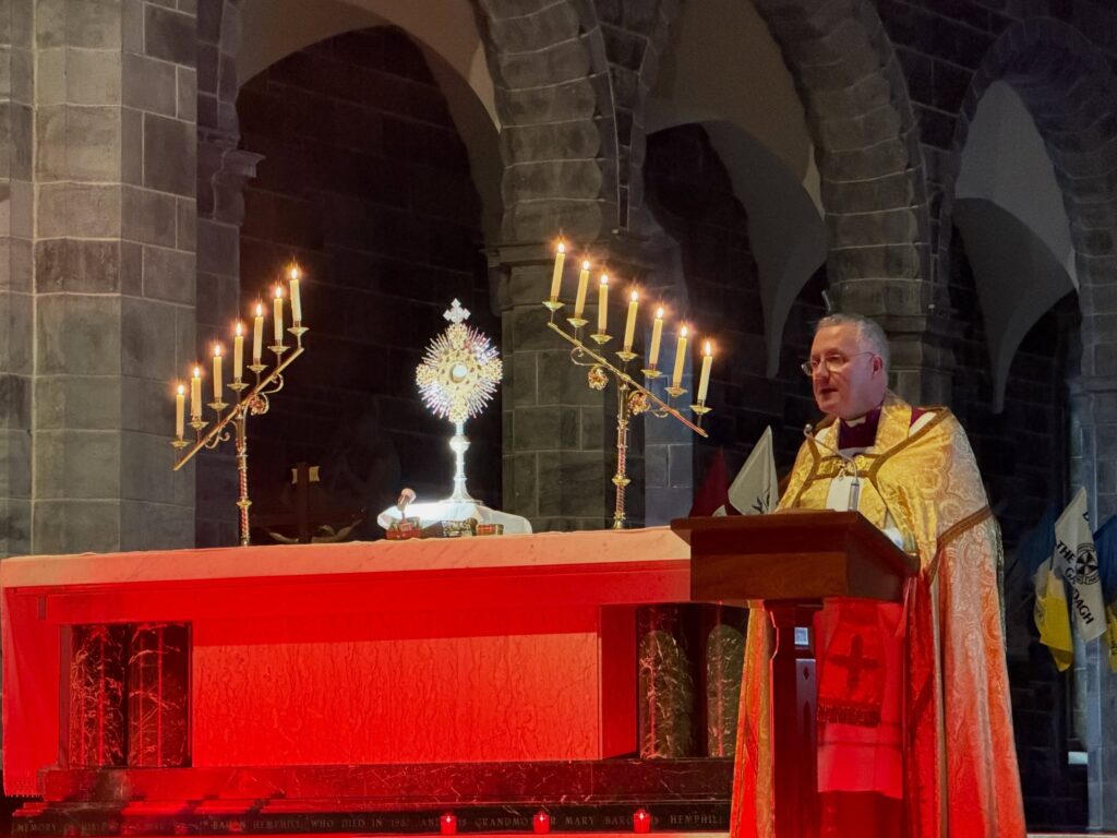 A priest in gold vestments speaks at a red-lit altar with candles and a monstrance in a stone church.