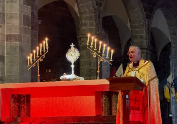 A priest in gold vestments speaks at a red-lit altar with candles and a monstrance in a stone church.