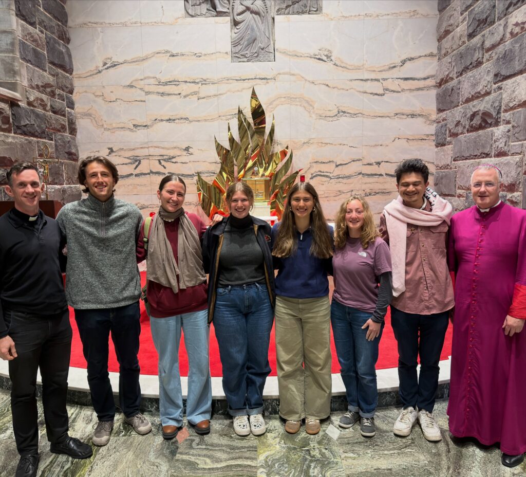 Seven young adults and two clergy stand smiling together inside a church, in front of a gold, flame-like altar piece.