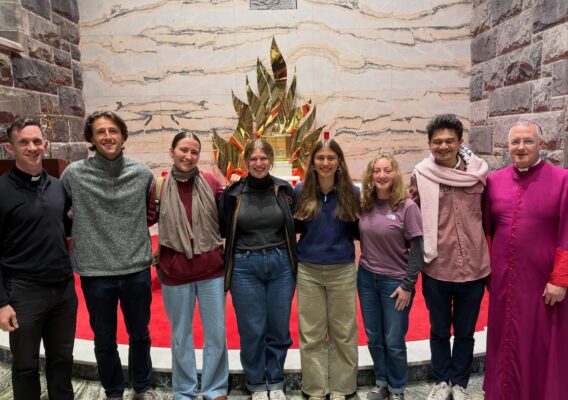 Seven young adults and two clergy stand smiling together inside a church, in front of a gold, flame-like altar piece.
