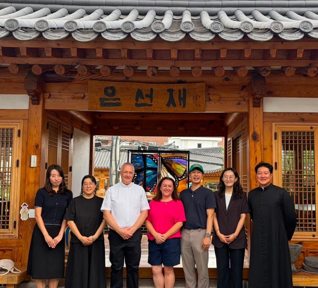 Seven people stand smiling in front of a traditional Korean building with a wooden sign and tiled roof.