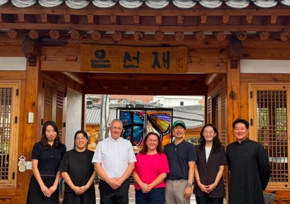 Seven people stand smiling in front of a traditional Korean building with a wooden sign and tiled roof.
