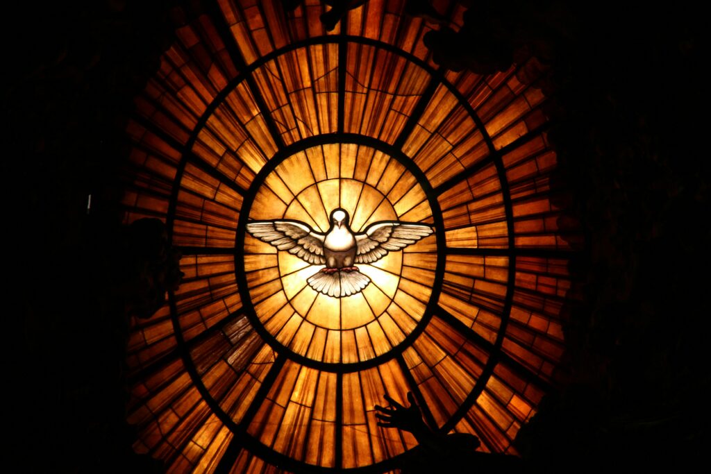Stained glass window of a white dove with outstretched wings, glowing with warm amber light in a circular pattern.