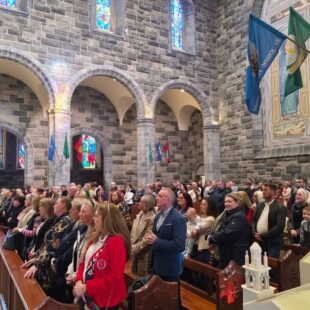 A congregation stands in a stone church with arched doorways, stained glass windows, and hanging flags.