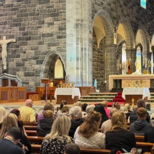 A man in a suit speaks into a microphone at the altar of a stone church filled with a seated congregation.