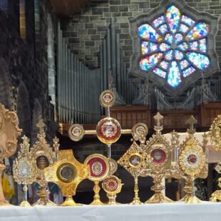 A collection of ornate gold religious reliquaries displayed on an altar in front of a stone wall and a stained-glass window.