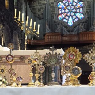 An altar covered with a collection of ornate golden reliquaries and a monstrance inside a stone church.